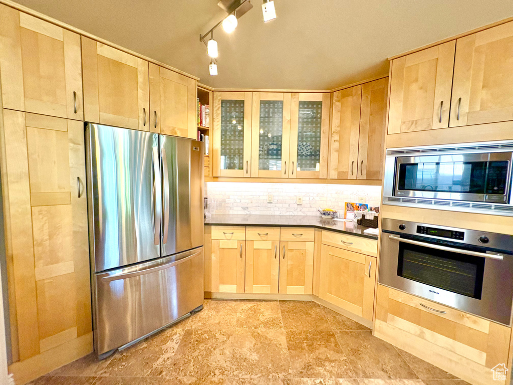 Kitchen with appliances with stainless steel finishes, light brown cabinetry, decorative backsplash, glass insert cabinets, and dark countertops