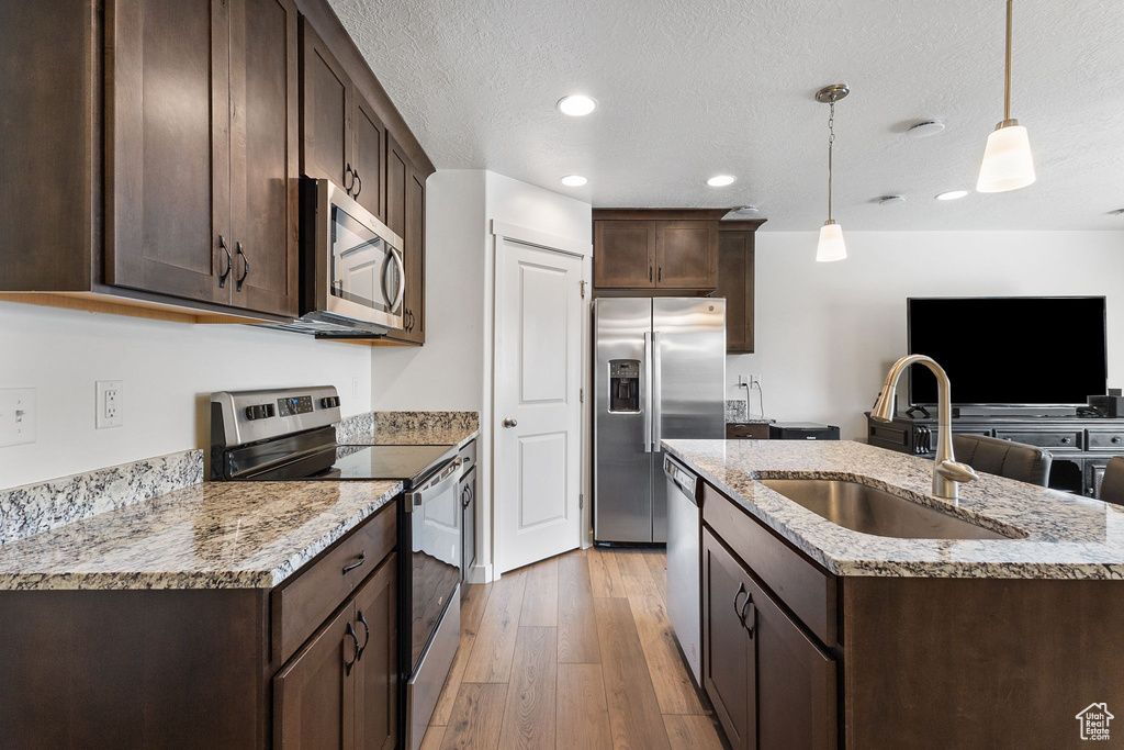 Kitchen featuring appliances with stainless steel finishes, dark brown cabinetry, light wood-type flooring, a textured ceiling, and an island with sink