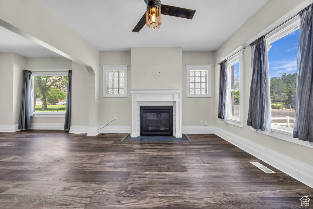 Unfurnished living room with a ceiling fan, plenty of natural light, a tiled fireplace, and dark wood finished floors