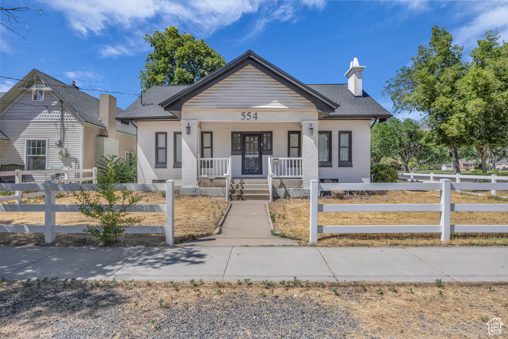 Bungalow-style home featuring covered porch, brick siding, a fenced front yard, a chimney, and a shingled roof