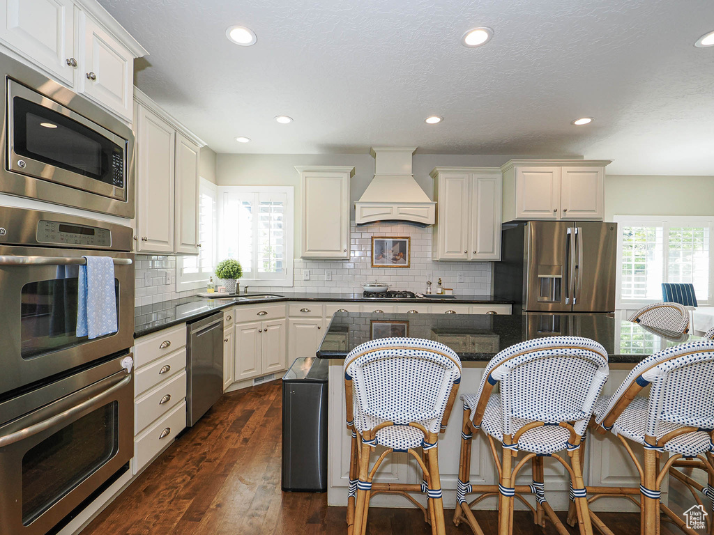 Kitchen with stainless steel appliances, custom range hood, dark countertops, a kitchen breakfast bar, and recessed lighting