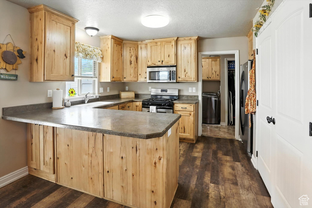 Kitchen with appliances with stainless steel finishes, a peninsula, dark wood finished floors, light brown cabinetry, and a textured ceiling