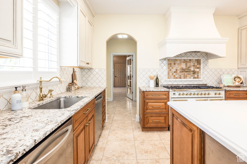 Kitchen featuring stainless steel dishwasher, premium range hood, brown cabinets, and light stone counters