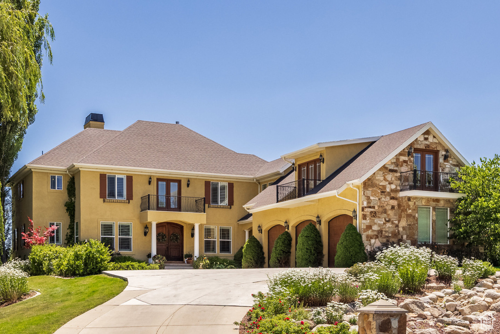 View of front of home with a balcony, french doors, concrete driveway, stone siding, and stucco siding