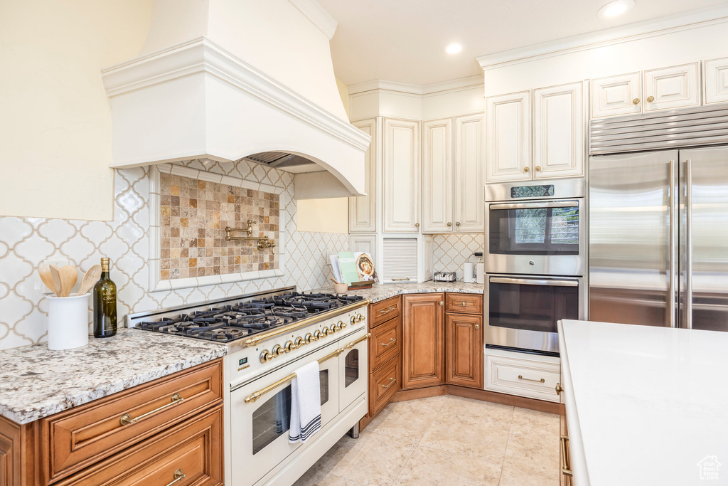 Kitchen featuring high end appliances, premium range hood, brown cabinetry, light stone counters, and recessed lighting