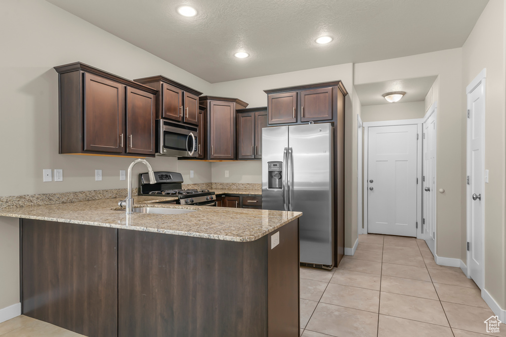 Kitchen featuring appliances with stainless steel finishes, a peninsula, dark brown cabinets, light tile patterned floors, and light stone countertops