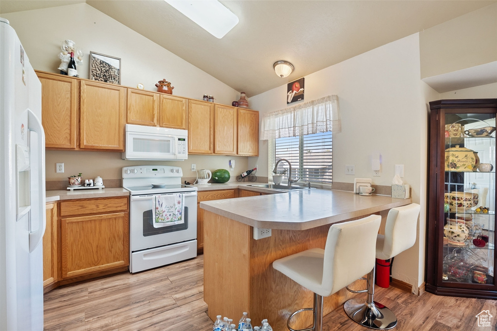 Kitchen with white appliances, a peninsula, vaulted ceiling, light wood-style flooring, and a kitchen breakfast bar