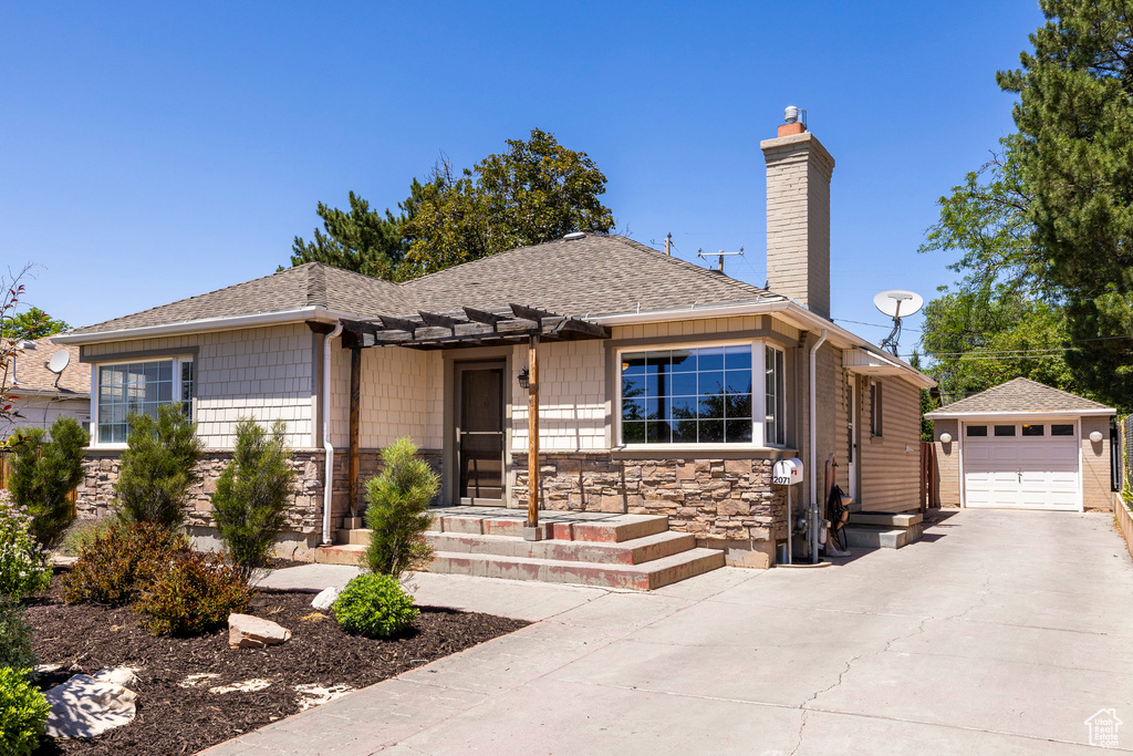 View of front of house featuring a chimney, concrete driveway, an outdoor structure, a shingled roof, and stone siding