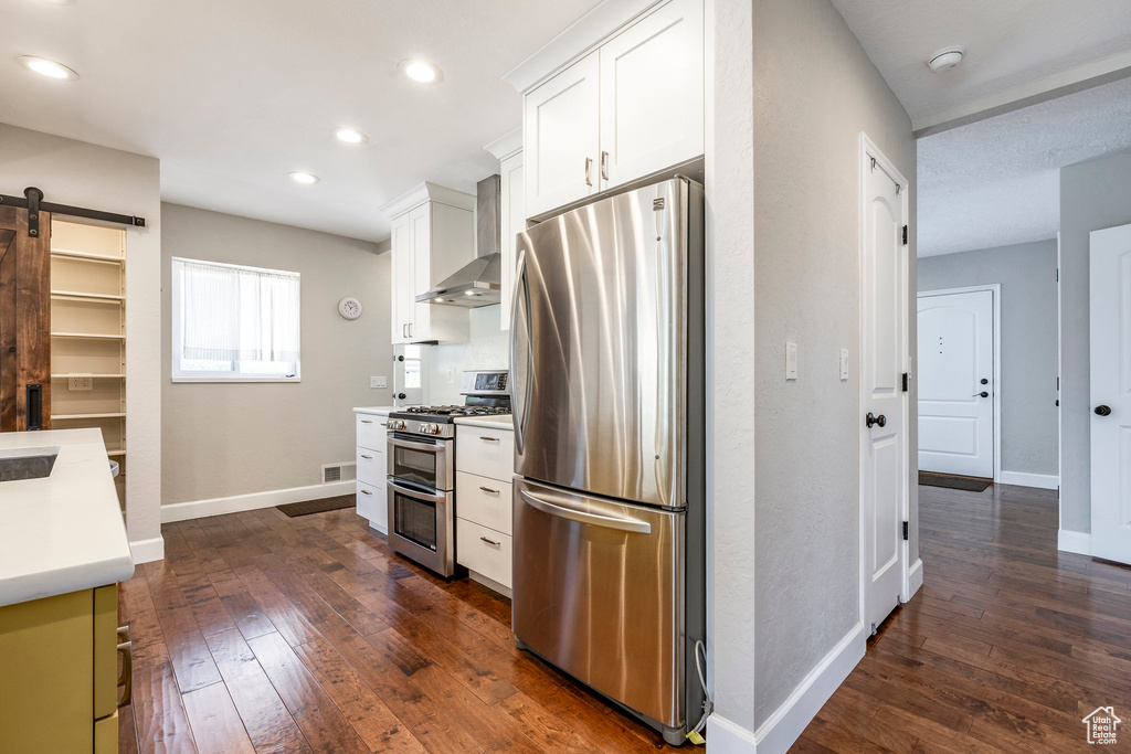 Kitchen featuring appliances with stainless steel finishes, wall chimney exhaust hood, a barn door, dark wood-type flooring, and white cabinetry
