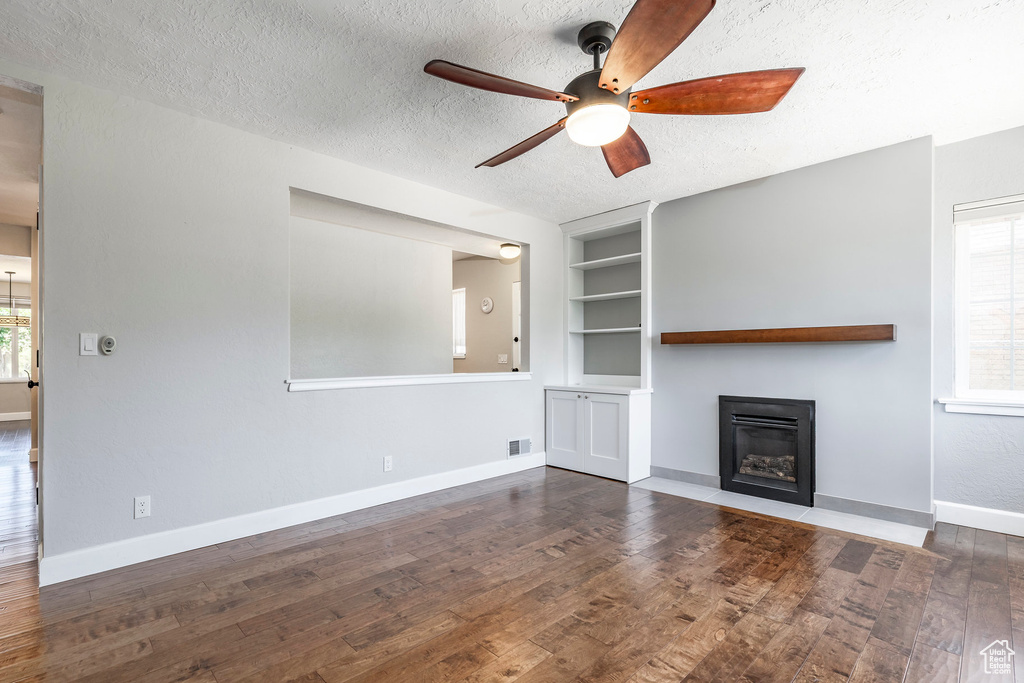 Unfurnished living room featuring a ceiling fan, a glass covered fireplace, a textured ceiling, built in shelves, and wood finished floors