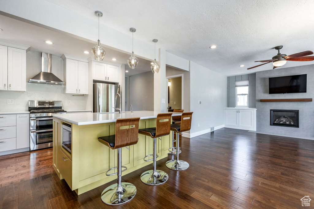 Kitchen with appliances with stainless steel finishes, wall chimney range hood, a kitchen island with sink, light countertops, and dark wood-style flooring