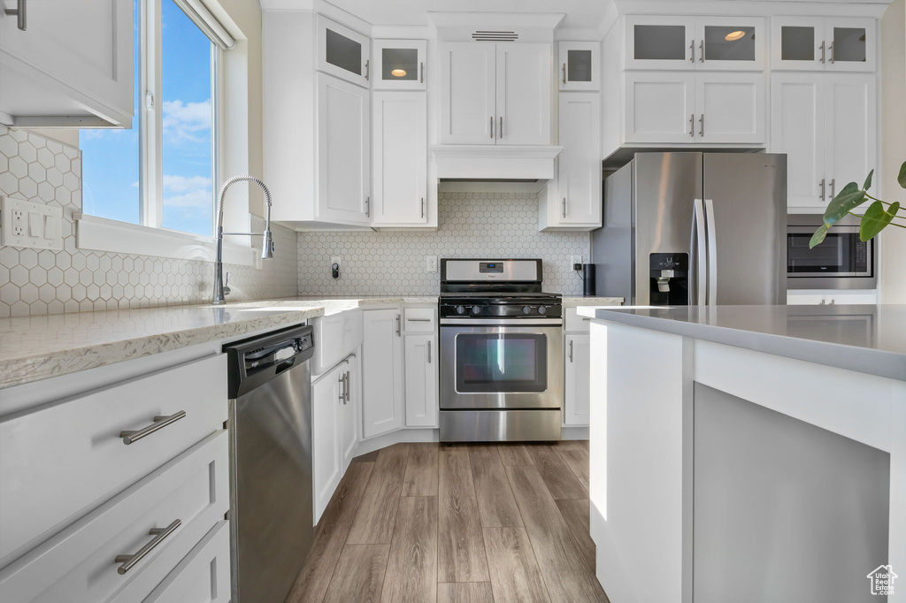 Kitchen with stainless steel appliances, light wood-style flooring, white cabinets, tasteful backsplash, and glass insert cabinets