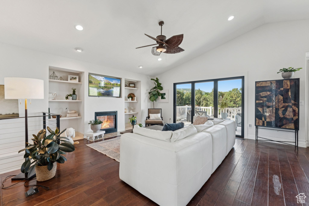 Living room with ceiling fan, vaulted ceiling, a glass covered fireplace, built in shelves, and dark wood-style floors