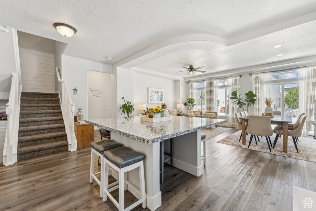 Kitchen with a textured ceiling, dark wood-type flooring, a kitchen bar, light stone counters, and a center island