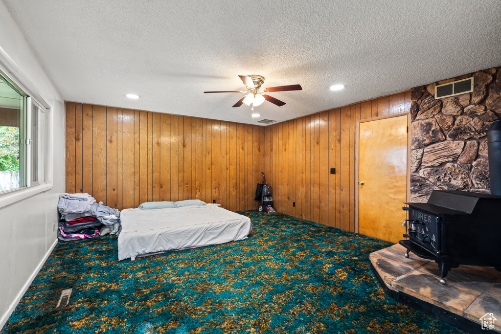Carpeted bedroom featuring a wood stove, a textured ceiling, wooden walls, recessed lighting, and a ceiling fan