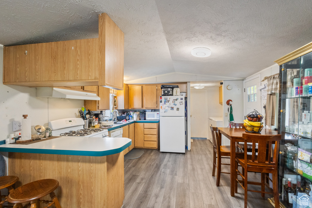 Kitchen featuring white appliances, under cabinet range hood, lofted ceiling, a peninsula, and light wood-style floors