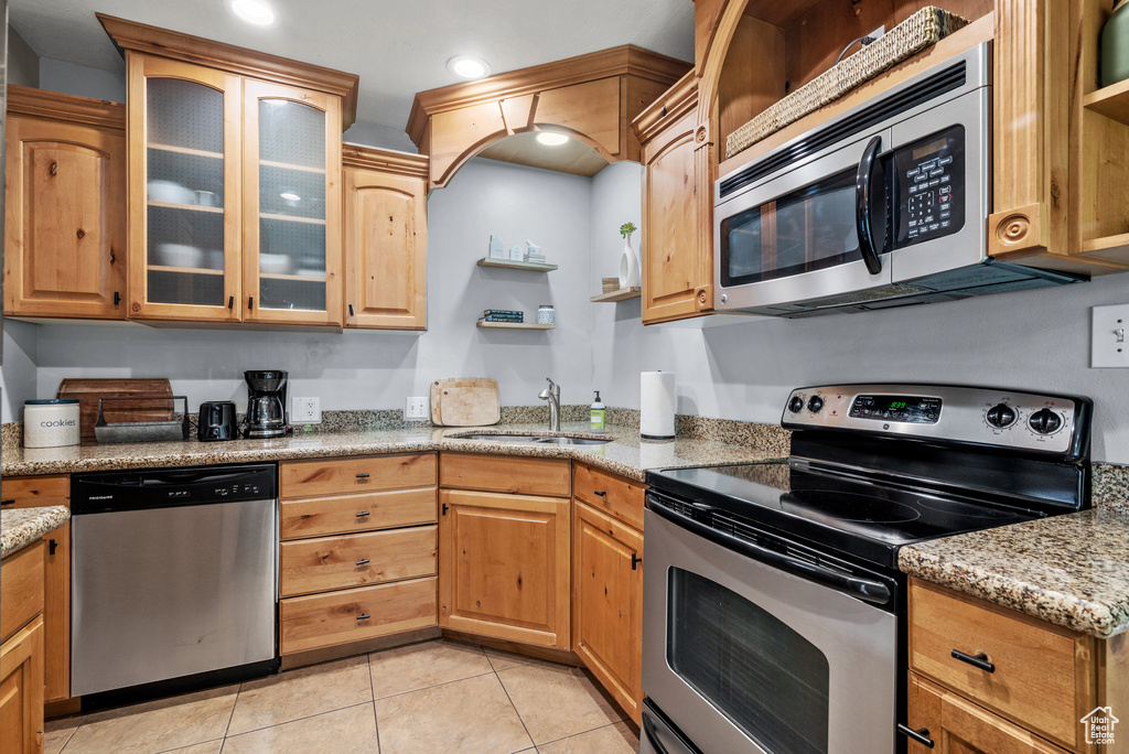 Kitchen with appliances with stainless steel finishes, open shelves, light tile patterned flooring, glass insert cabinets, and light stone counters