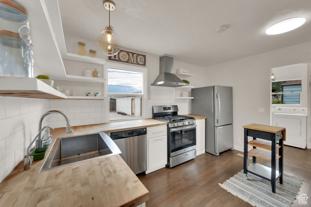 Kitchen featuring wall chimney exhaust hood, stainless steel appliances, open shelves, butcher block counters, and tasteful backsplash