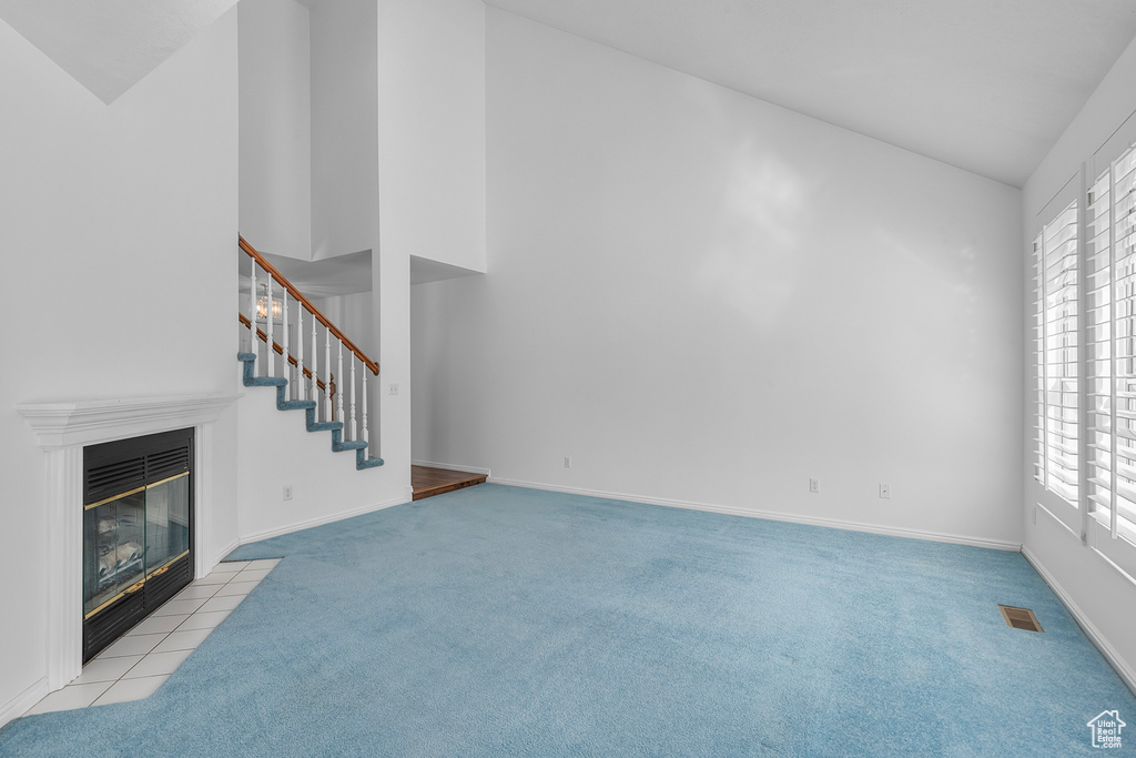 Unfurnished living room featuring light colored carpet, healthy amount of natural light, a tiled fireplace, stairs, and high vaulted ceiling