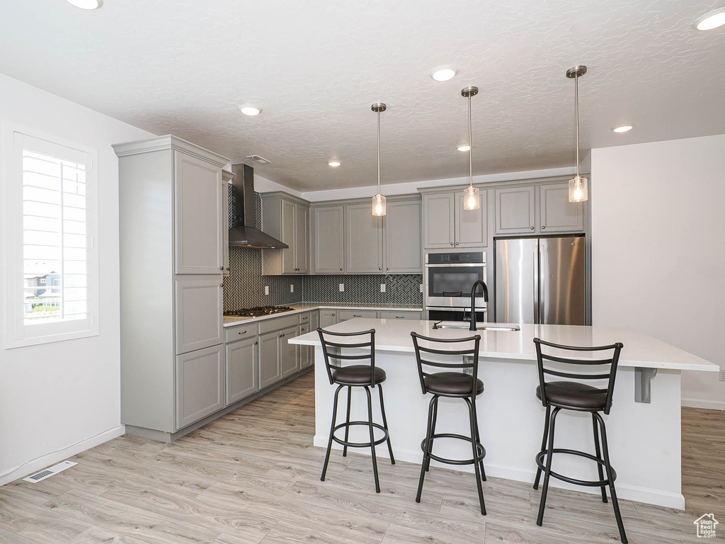 Kitchen with stainless steel appliances, gray cabinetry, wall chimney exhaust hood, light countertops, and a center island with sink