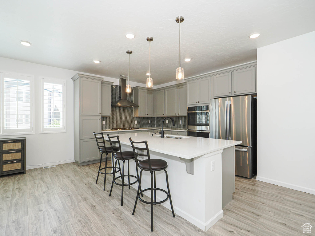 Kitchen with gray cabinetry, wall chimney range hood, appliances with stainless steel finishes, light wood-style floors, and recessed lighting