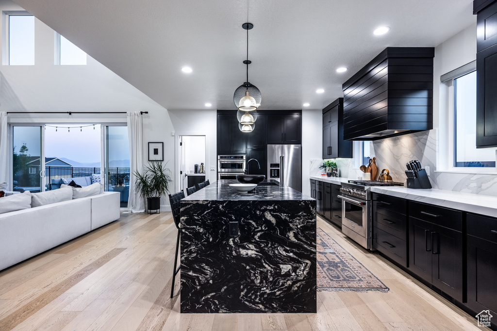Kitchen with appliances with stainless steel finishes, dark cabinetry, wall chimney exhaust hood, light stone countertops, and backsplash