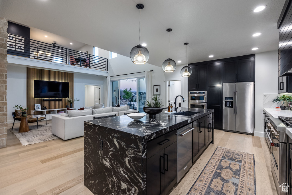 Kitchen with high quality appliances, dark cabinetry, light wood-type flooring, an island with sink, and a towering ceiling