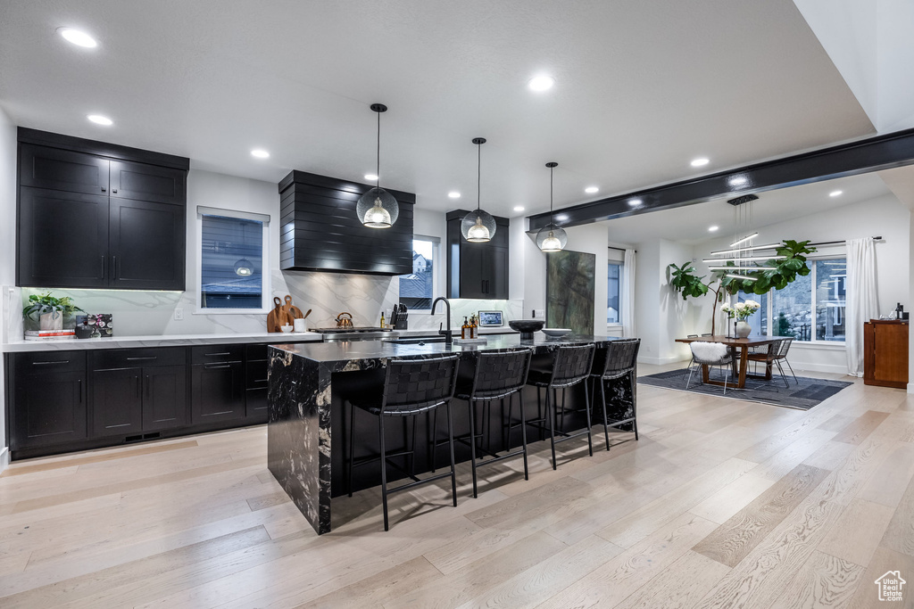 Kitchen with wall chimney range hood, dark cabinets, backsplash, a kitchen breakfast bar, and light wood-type flooring