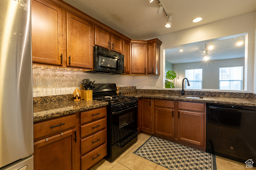 Kitchen with black appliances, track lighting, brown cabinetry, dark stone counters, and light tile patterned floors