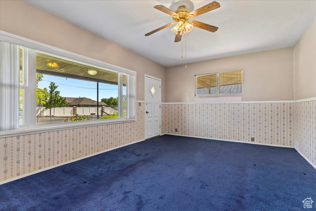 Empty room featuring wallpapered walls, wainscoting, dark colored carpet, and ceiling fan