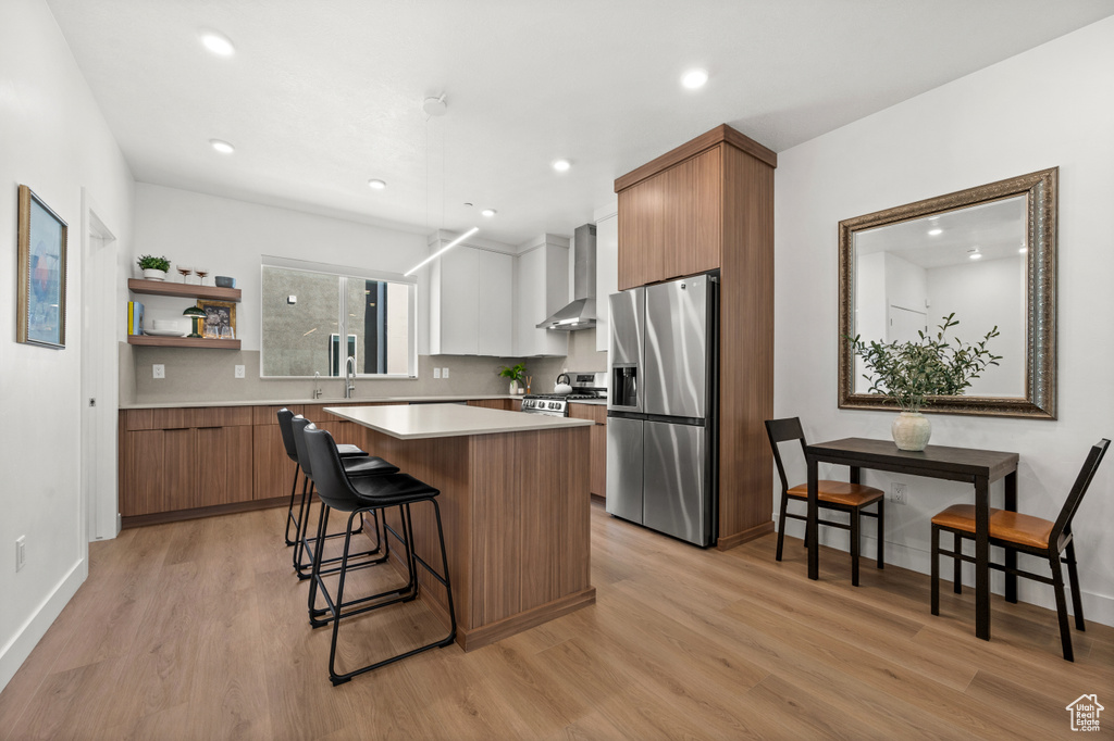 Kitchen with appliances with stainless steel finishes, wall chimney exhaust hood, a center island, a breakfast bar area, and light wood-style floors