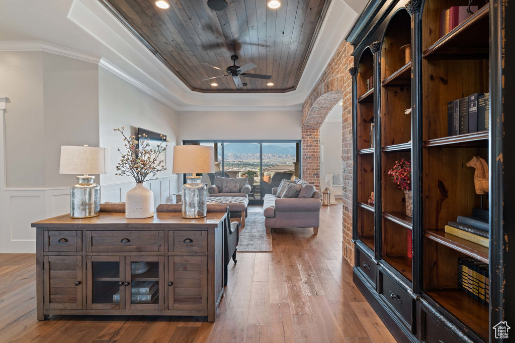Living room with a raised ceiling, arched walkways, light wood finished floors, a ceiling fan, and a wainscoted wall
