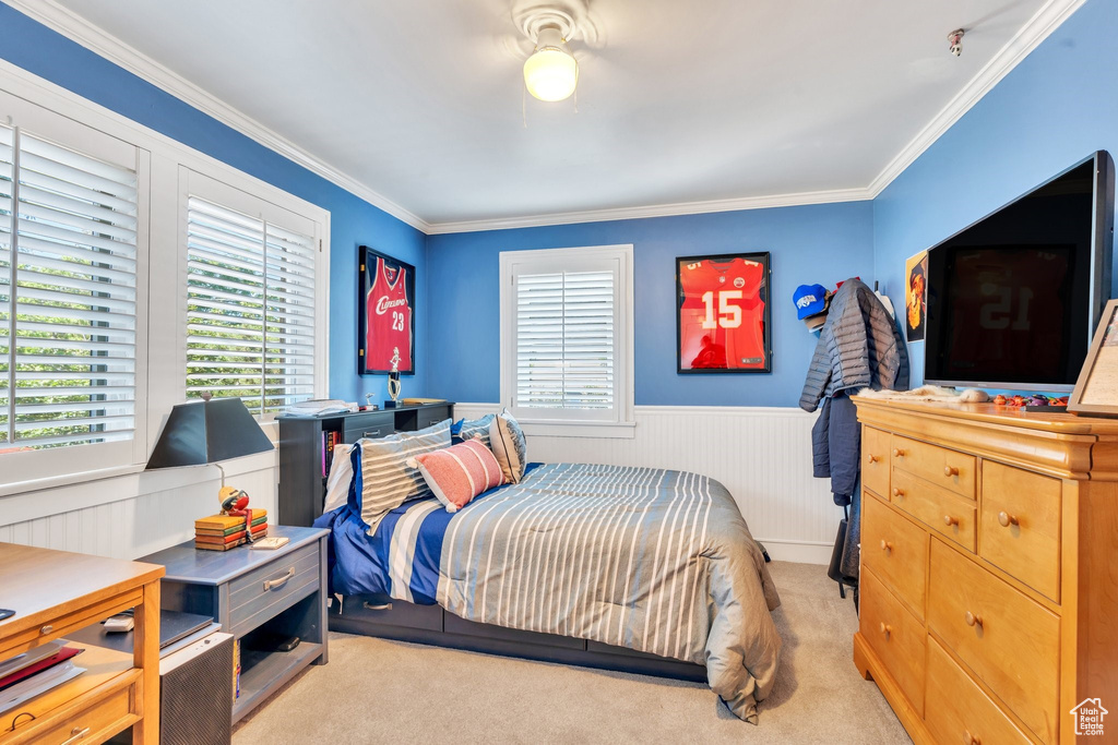 Bedroom featuring multiple windows, a wainscoted wall, light colored carpet, crown molding, and ceiling fan