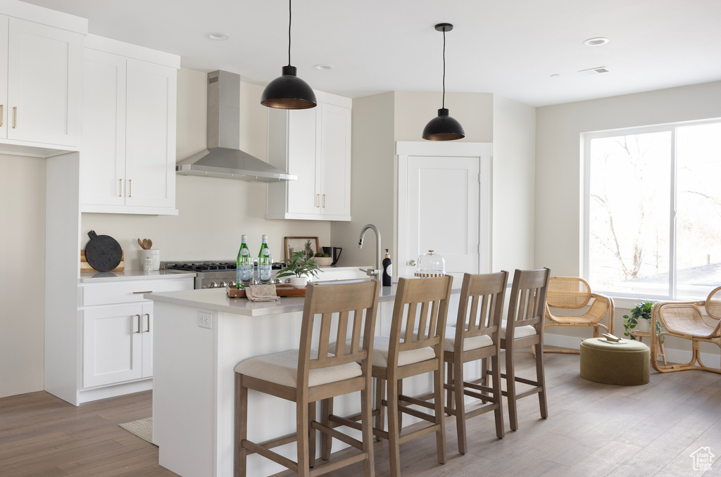 Kitchen featuring wall chimney exhaust hood, light wood-style floors, light countertops, an island with sink, and white cabinetry
