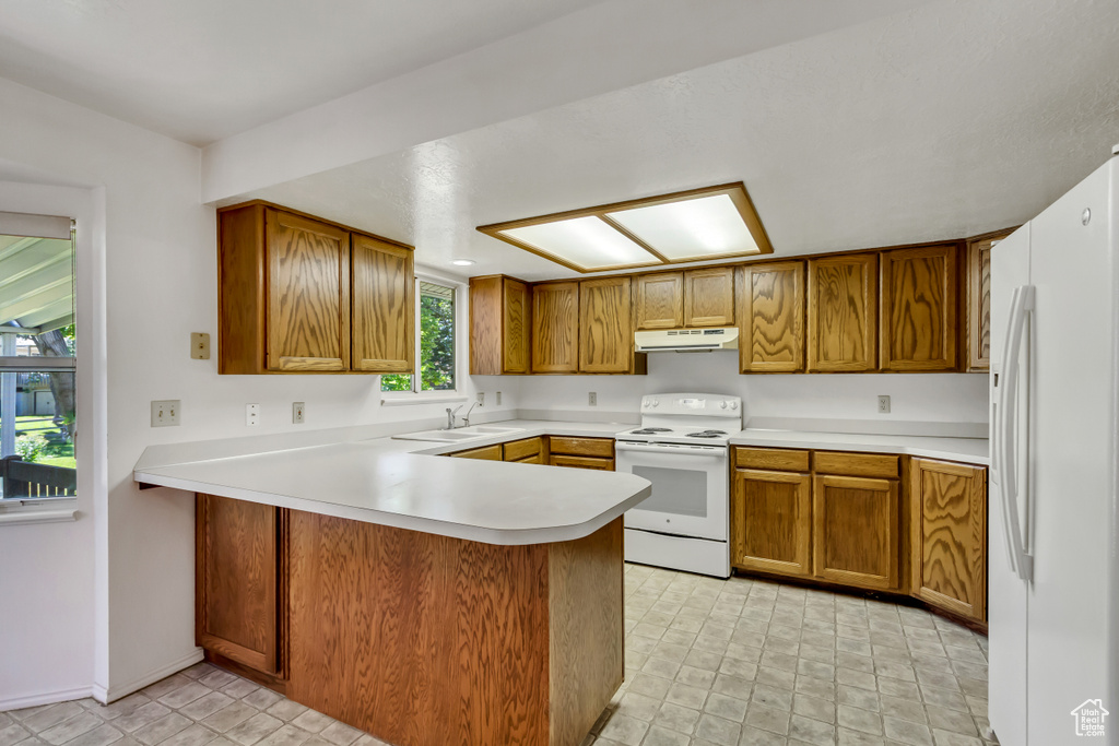 Kitchen featuring white appliances, under cabinet range hood, a peninsula, brown cabinets, and light countertops