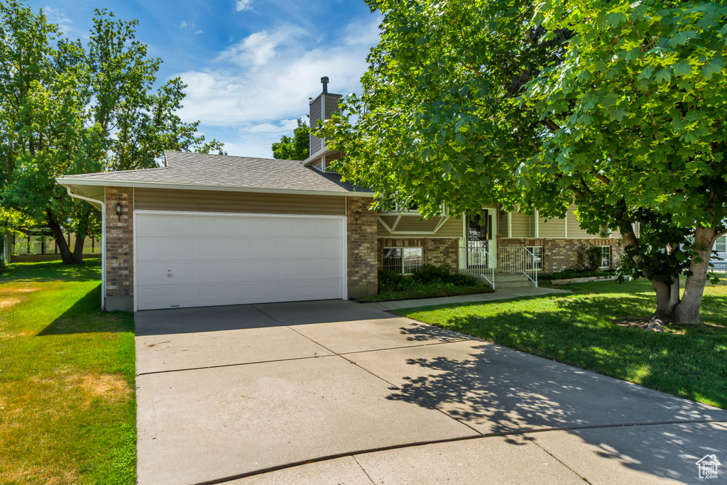 View of property hidden behind natural elements featuring a front lawn, brick siding, concrete driveway, a garage, and a chimney