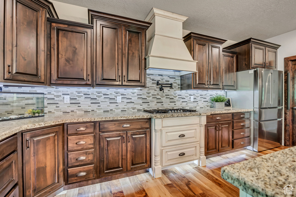 Kitchen featuring stainless steel appliances, custom range hood, light wood-style flooring, dark brown cabinets, and decorative backsplash