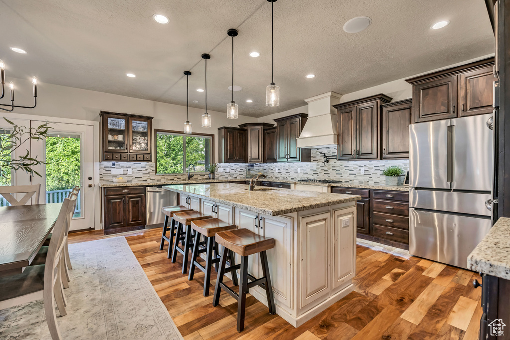 Kitchen with stainless steel appliances, premium range hood, a center island with sink, dark brown cabinetry, and a textured ceiling