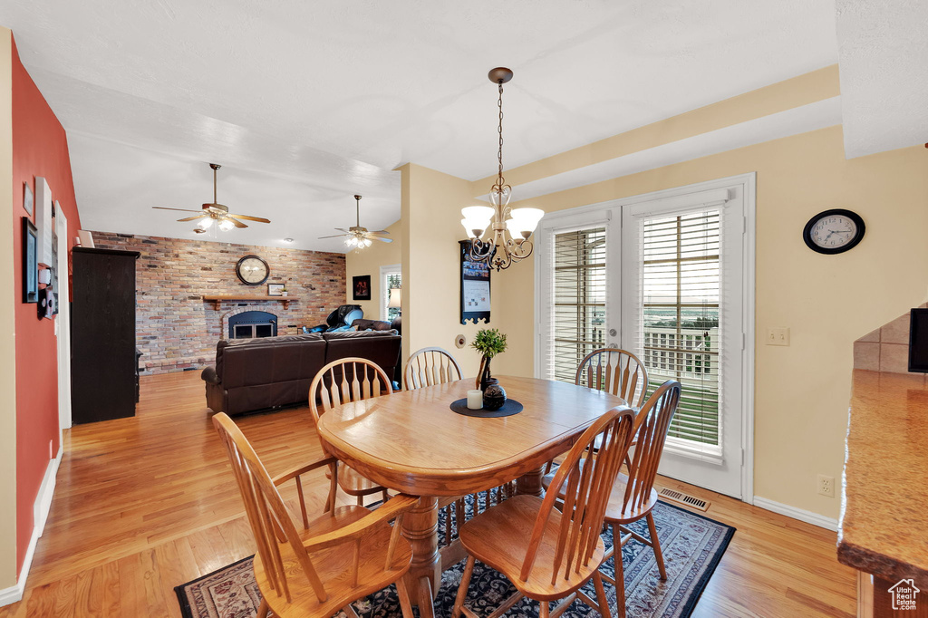 Dining room with light wood-style flooring, a ceiling fan, a fireplace, brick wall, and a chandelier