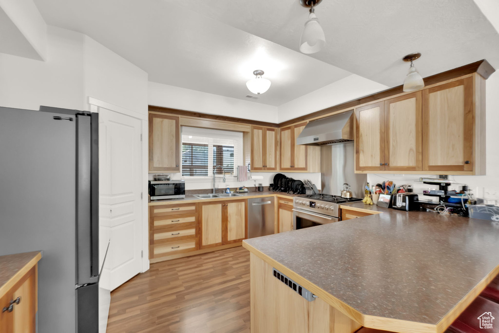 Kitchen featuring stainless steel appliances, wall chimney exhaust hood, light brown cabinetry, light wood finished floors, and a peninsula