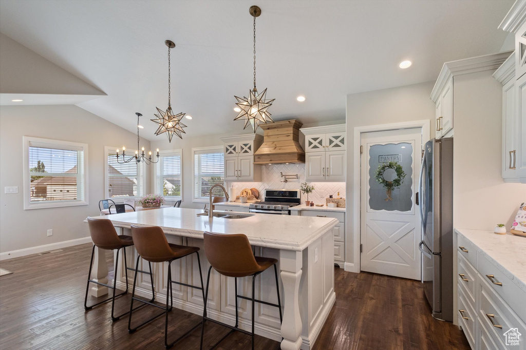 Kitchen with premium range hood, appliances with stainless steel finishes, a chandelier, tasteful backsplash, and a kitchen island with sink