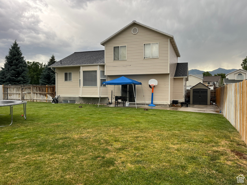 Back of house with a trampoline, a storage shed, a fenced backyard, a patio area, and roof with shingles