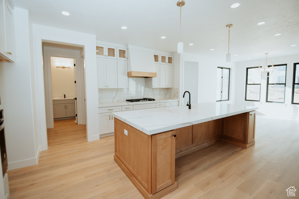 Kitchen featuring premium range hood, light wood-type flooring, glass insert cabinets, an island with sink, and light stone counters