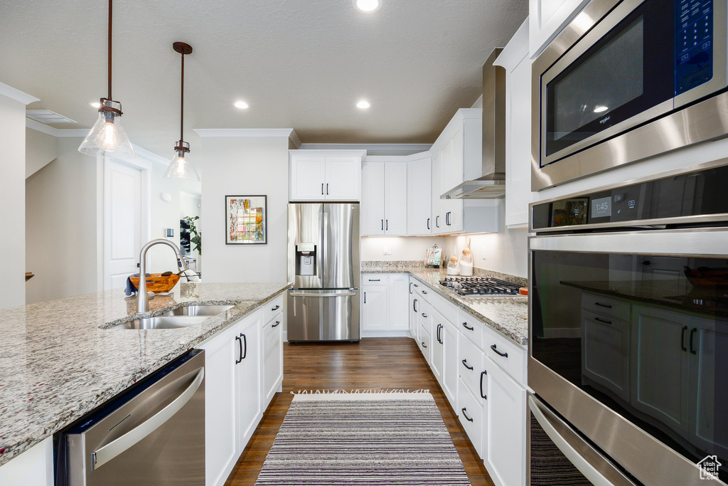 Kitchen with appliances with stainless steel finishes, wall chimney range hood, dark wood-type flooring, light stone counters, and hanging light fixtures