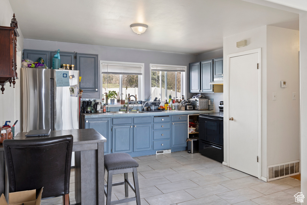 Kitchen with black / electric stove, freestanding refrigerator, under cabinet range hood, blue cabinets, and light countertops
