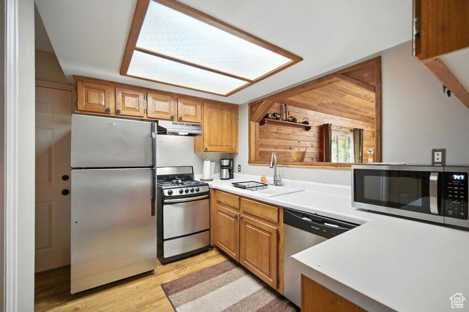 Kitchen featuring stainless steel appliances, under cabinet range hood, light wood finished floors, light countertops, and brown cabinets