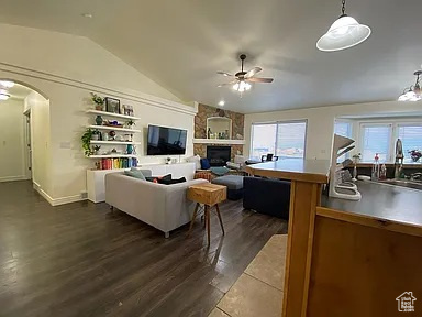 Living room with a ceiling fan, vaulted ceiling, arched walkways, dark wood-style floors, and a stone fireplace