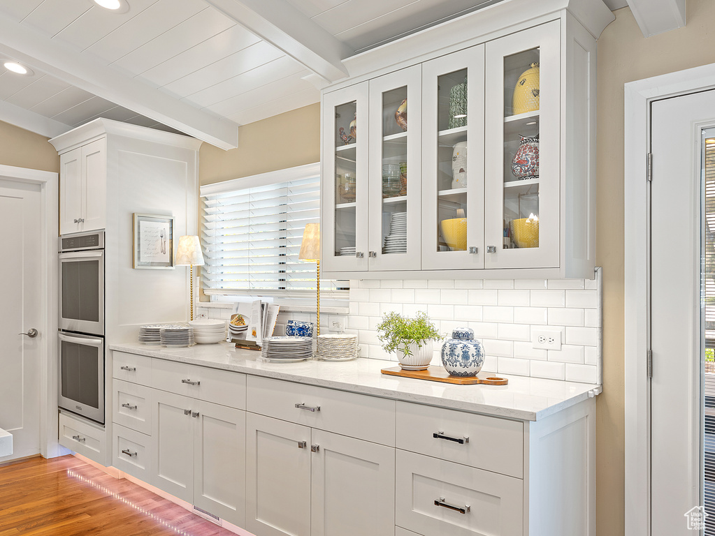 Kitchen featuring double oven, beamed ceiling, backsplash, white cabinetry, and glass insert cabinets