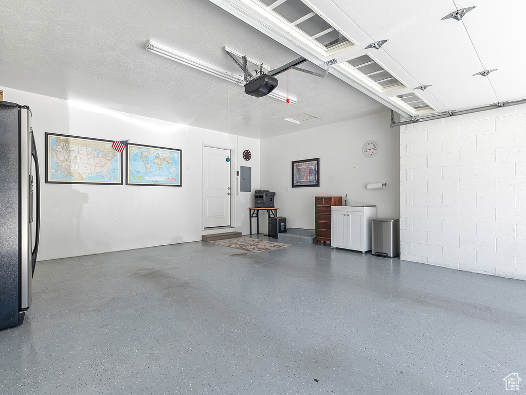 Garage with freestanding refrigerator, electric panel, a garage door opener, and concrete block wall