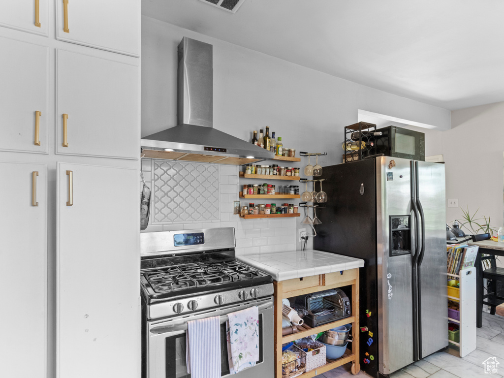 Kitchen featuring wall chimney exhaust hood, appliances with stainless steel finishes, tasteful backsplash, tile countertops, and white cabinetry